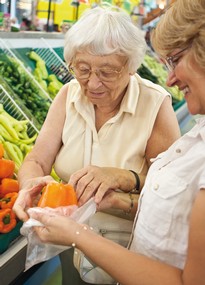Eine jüngere Frau hilft einer älteren Frau beim Gemüseregal, ein Stück Kürbis in einer Plastiktüte zu verstauen. 