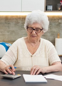 Eine ältere Frau mit markanter schwarzer Brille sitzt konzentriert am Küchentisch mit einem Taschenrechner und Schreibheft. 