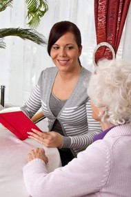 Eine jüngere Frau liest einer alten Person aus einem Buch mit rotem Einband vor. 