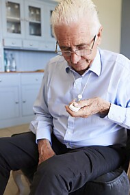 Ein älterer Mann mit Brille in helblauem Hemd und dunkler Hose drückt auf einen weissen Notrufknopf, den er mit einer an einer Kette um den Hals trägt. 